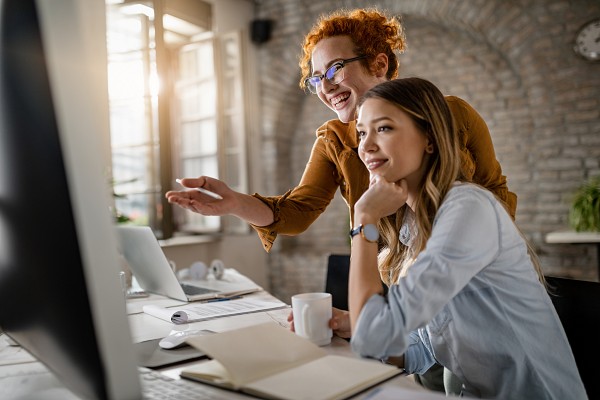 Young happy businesswomen cooperating while using desktop PC in | Playmotiv - Gamificación para empresas como elegir agencia de gamificacion